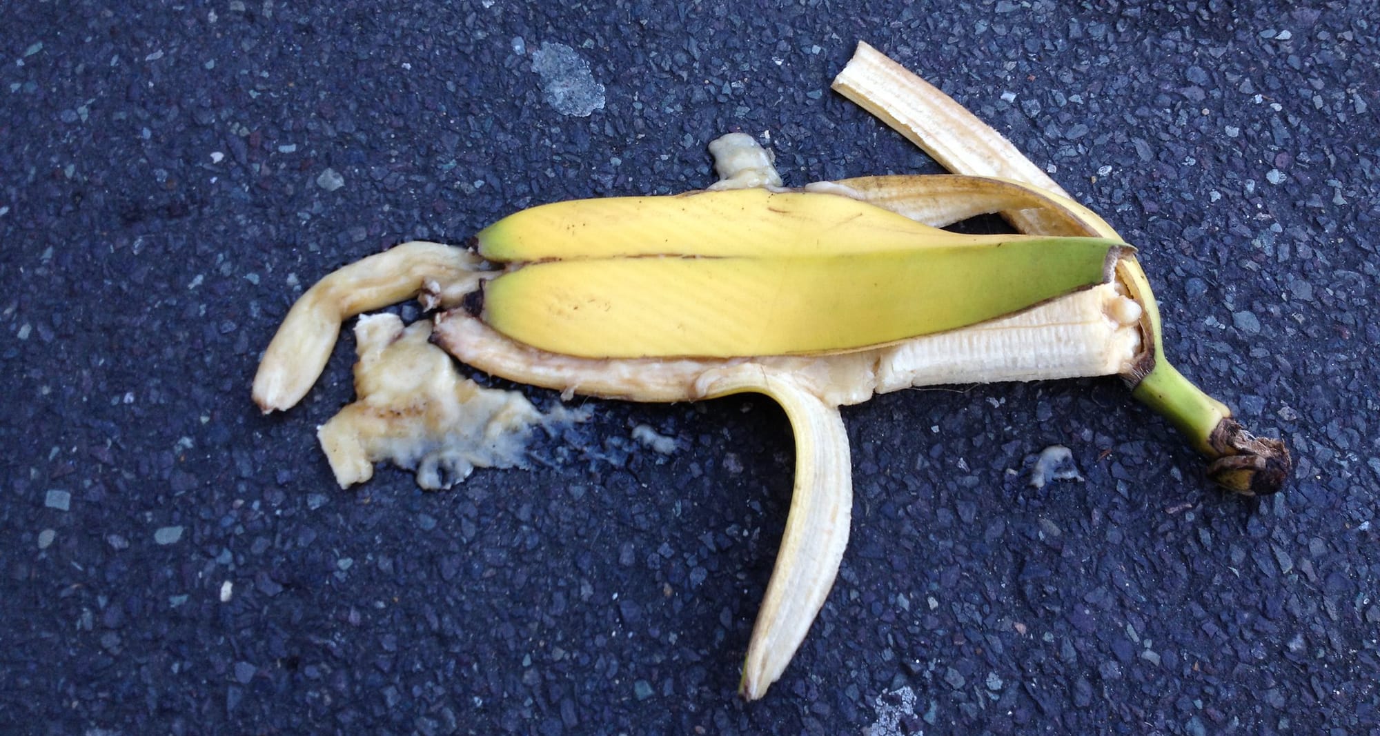 Close-up of a squashed, overripe yellow banana peel discarded on dark, textured asphalt. The fruit is partially mashed and oozing onto the pavement, creating a messy, flattened shape.